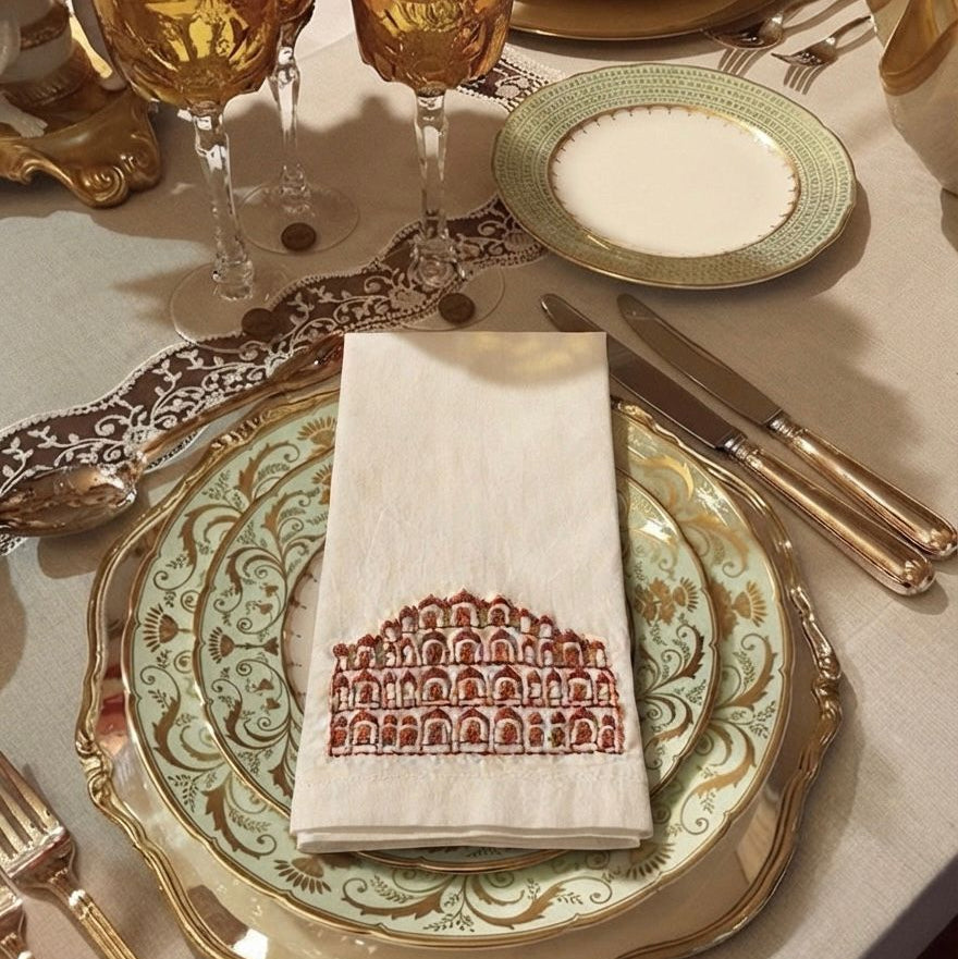 A styled table setting featuring a folded cream linen napkin with a red architectural arch pattern, layered over ornate gold and green plates on a beige tablecloth, alongside gold goblets and cutlery.
