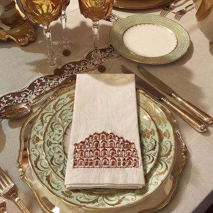 A styled table setting featuring a folded cream linen napkin with a red architectural arch pattern, layered over ornate gold and green plates on a beige tablecloth, alongside gold goblets and cutlery.