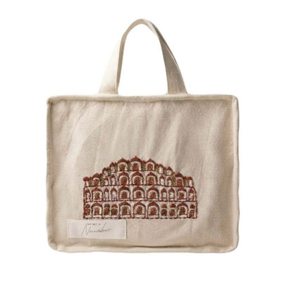 A beige canvas tote bag with a brown architectural illustration of a palace on the front, featuring two top handles and a small white brand label near the bottom left.
