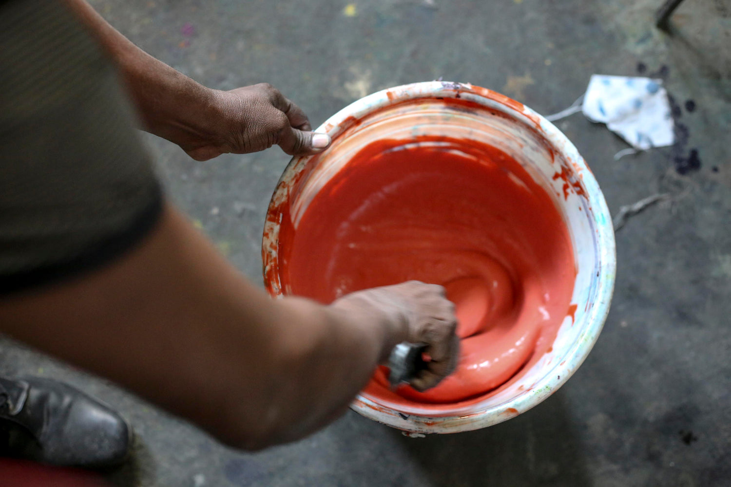 Person stirring red paint in a bucket with a brush on a concrete floor.